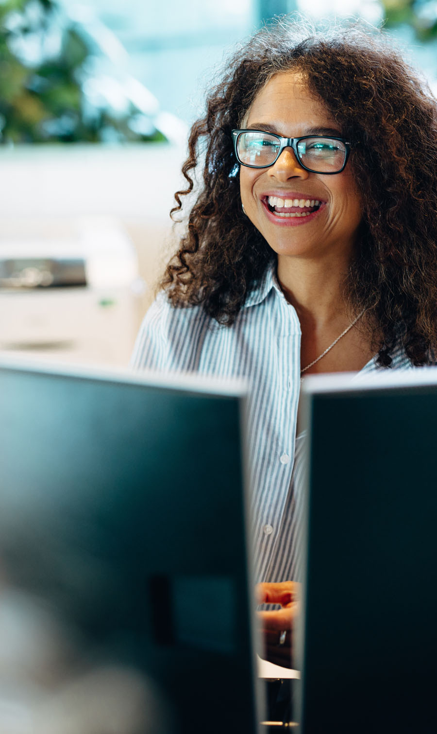 Image of a women at a desk behind two monitors wearing glasses and smiling.