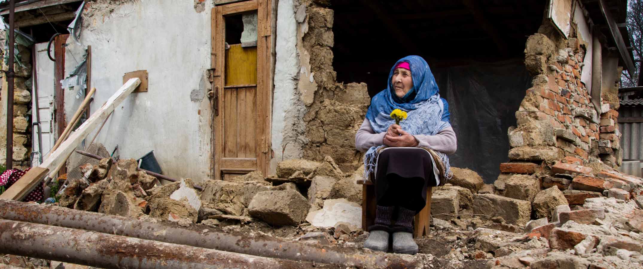 Photo shows an elderly woman holding flowers, sitting amongst a destroyed building.